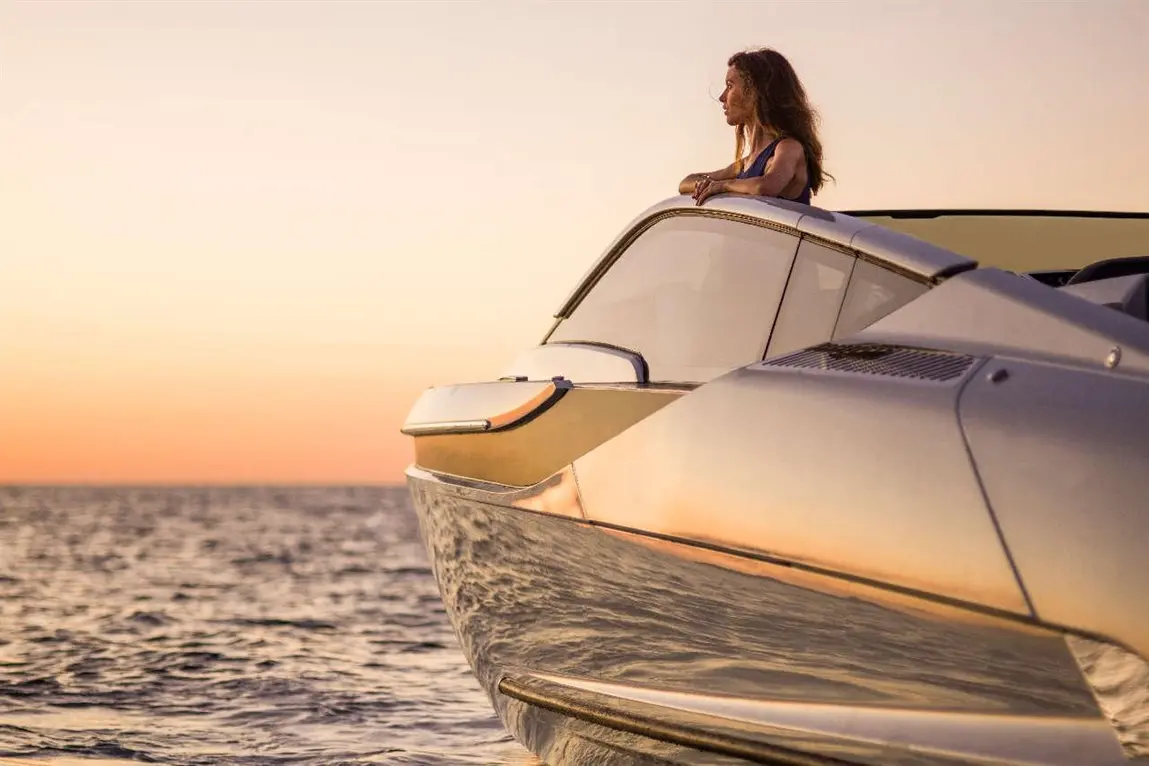 Woman in a blue dress leans on the bow of a sleek yacht, gazing at the sunset over calm ocean waters.